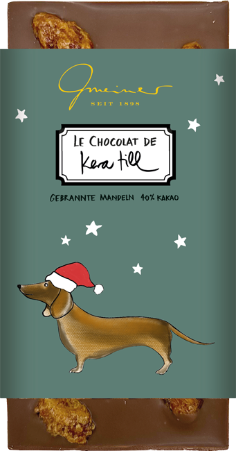 Gmeiner Kera Till Christmas Dog Schokolade mit Mandelstücken in dunkelgrüner Verpackung, brauner Dackel mit roter Zipfelmütze.